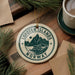 A circular ornament with the text "Lofoten Islands Norway" on it, surrounded by pine branches and a cup of coffee on a wooden surface.