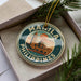 A circular ornament with the text "MANILA PHILIPPINES" and an illustration of a building, placed in a box with pine branches.