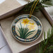 A decorative ceramic ornament with a floral design, featuring yellow flowers and green leaves, is displayed in a box with pine branches.