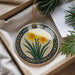 A decorative ceramic ornament with a floral design, featuring yellow daffodils, is displayed in a box with pine branches and a white card.