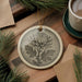 a round ceramic ornament with a detailed chrysanthemum design, placed on a wooden surface alongside a cup of coffee and some pine branches.