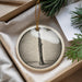 A circular ornament featuring a black and white illustration of a clarinet is displayed in a box, surrounded by pine branches.