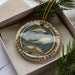 A decorative ornament in a box, featuring a design with two fish and a castle or building.