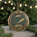 A decorative ornament featuring a fish and a bridge, with the word "PIGES" written around the edge. The ornament is hanging from a string, and it is placed on a surface with pine branches in the background.