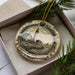 A decorative ornament featuring a lighthouse and a coastal scene, placed in a box with pine branches.