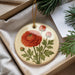 A round ornament with a red flower and green leaves is displayed in a box, surrounded by pine branches.