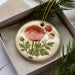 A decorative ornament featuring a large red flower surrounded by green leaves, placed in a box with pine branches.