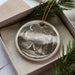 A ceramic ornament depicting a camping scene with a tent, campfire, and mountains in the background, hanging from a string.