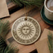 a decorative ceramic ornament with a sun design, placed on a wooden surface alongside a cup of coffee and some pine branches.