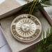 A decorative ceramic ornament with a sun design is displayed in a box, surrounded by pine branches.