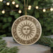A round ornament with a sun design is hanging from a string, surrounded by pine branches and twinkling lights in the background.