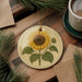 a round ceramic ornament with a sunflower design, placed on a wooden surface alongside a cup of coffee and some pine branches.