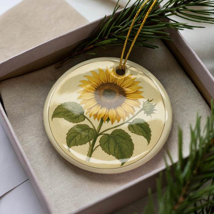 A round ornament with a sunflower design is placed inside a box, surrounded by pine branches.