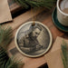 a ceramic ornament depicting a man engrossed in his work, surrounded by pine branches and a cup of coffee.