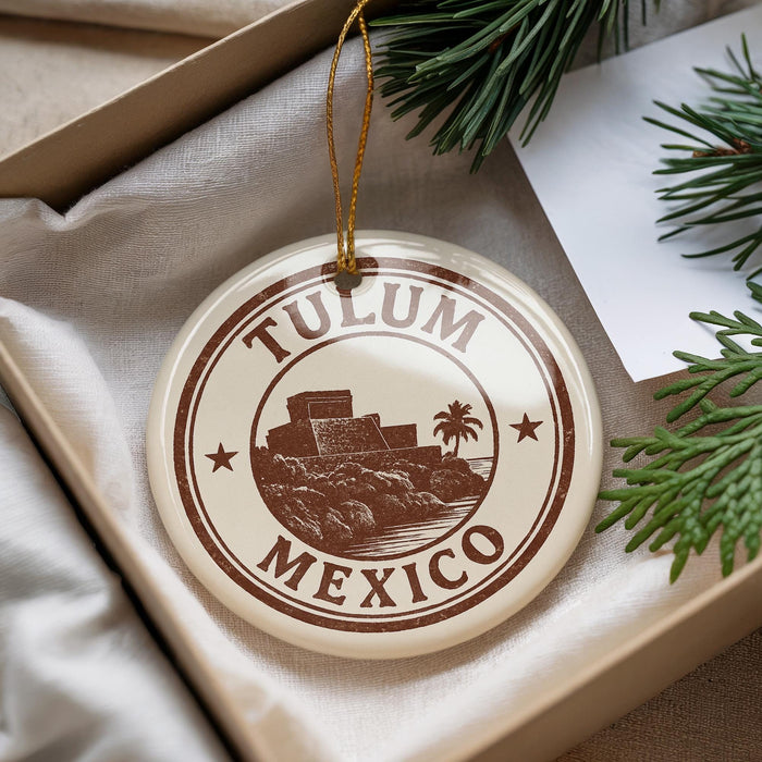 A circular ornament with the text "TULUM MEXICO" printed on it, surrounded by a border and a palm tree illustration. The ornament is hanging from a string, and there are pine branches visible in the background.