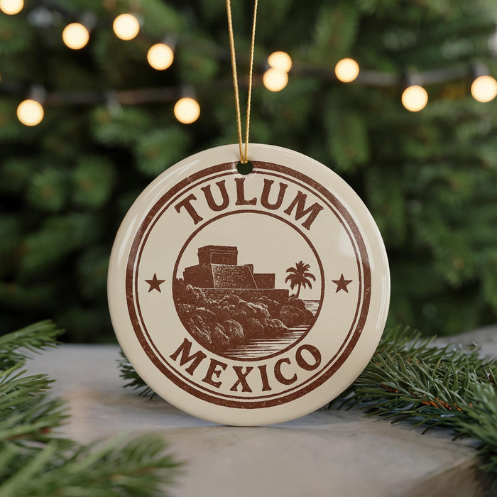 A circular ornament with the text "TULUM MEXICO" and an illustration of a landmark, possibly a castle or fort, is hanging from a string. The ornament is placed on a surface with pine branches in the background.