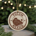A circular ornament with the text "TULUM MEXICO" and an illustration of a landmark, possibly a castle or fort, is hanging from a string. The ornament is placed on a surface with pine branches in the background.