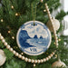 A Christmas tree decorated with a blue and white ornament featuring a coastal scene, surrounded by ornaments and lights.