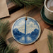 A decorative ceramic ornament featuring the Washington Monument, a tall obelisk, is displayed on a wooden surface alongside a cup of coffee and some pine branches.