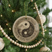 A decorative ornament with a yin and yang symbol is hanging on a Christmas tree, surrounded by other ornaments and lights.