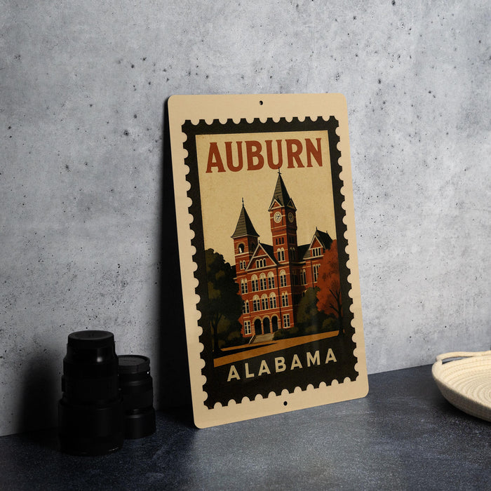 A metal sign with a stamp-like design, featuring a building and the text "AUBURN ALABAMA" on it, placed on a table next to some black camera lenses.