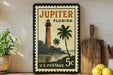 A vintage postage stamp from Jupiter, Florida, featuring a lighthouse and palm trees, is displayed on a shelf next to a wooden cutting board and a potted plant.