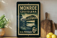 A vintage-style postage stamp from Monroe, Louisiana, featuring a bridge and water, displayed on a shelf next to a wooden cutting board and some lemons.