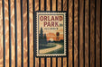 A vintage-style postage stamp depicting a scenic landscape with a winding road, trees, and a water tower, mounted on a wooden wall.