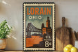 A vintage-style postage stamp from Ohio, featuring an illustration of the Ohio State Bridge, is displayed on a shelf next to a wooden cutting board and some lemons.