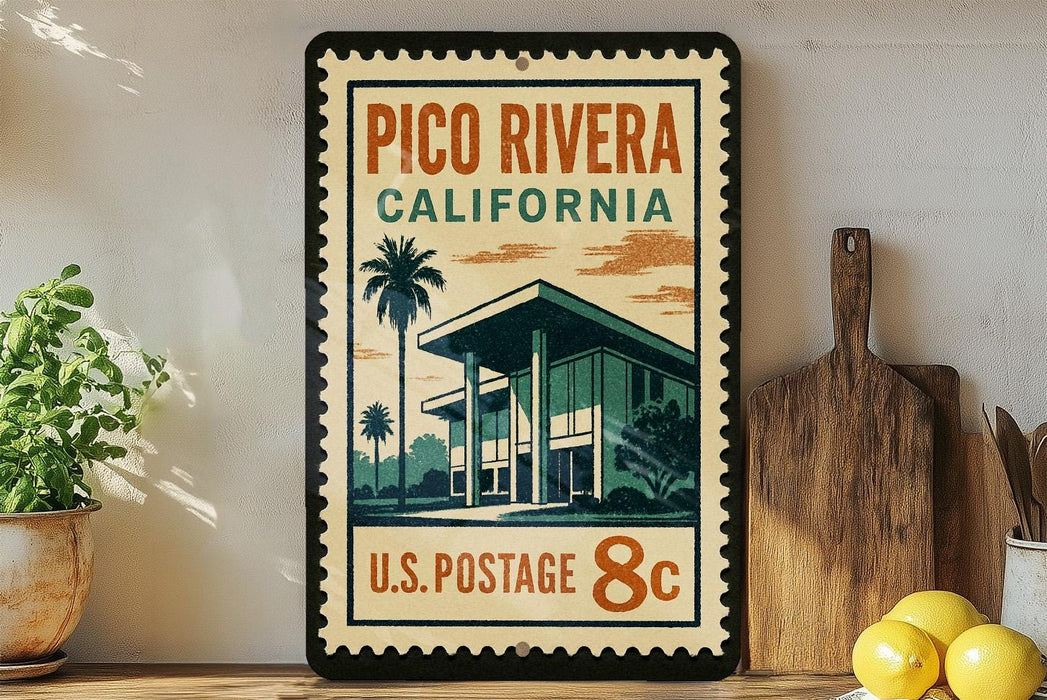 A vintage-style postage stamp featuring a building and palm trees, with the text "PICO RIVERA CALIFORNIA U.S. POSTAGE 8c" printed on it. The stamp is displayed on a shelf next to a wooden cutting board and a potted plant.
