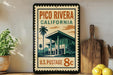 A vintage-style postage stamp featuring a building and palm trees, with the text "PICO RIVERA CALIFORNIA U.S. POSTAGE 8c" printed on it. The stamp is displayed on a shelf next to a wooden cutting board and a potted plant.