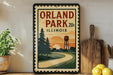 A vintage-style sign for Orland Park, Illinois, featuring a scenic landscape with a winding road, pine trees, and a water tower.