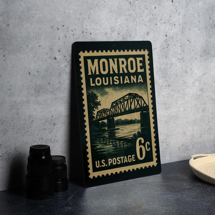 A vintage-style postage stamp from Monroe, Louisiana, featuring a bridge and water, placed on a countertop next to some black camera lenses.