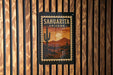 A framed poster of a desert landscape with a cactus and mountains in the background, mounted on a wooden wall.