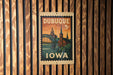 A vintage postage stamp from Dubuque, Iowa, featuring an illustration of a bridge and a church, mounted on a wooden wall.