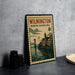 A metal sign with a vintage design depicting a cityscape and a bridge, placed on a table next to some black camera lenses.