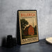 A vintage-style stamp depicting a red brick house with a church steeple, placed on a countertop next to some black camera lenses.