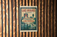 A vintage-style metal sign that reads "Welcome to Fabulous Las Vegas Nevada" and features a postage stamp design, mounted on a wooden wall.