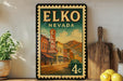 A vintage-style poster of Elko, Nevada, featuring a street scene with buildings, a hotel sign, and a car. The poster is framed and displayed on a shelf, with a wooden cutting board and a potted plant nearby.