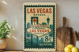A vintage-style metal sign that reads "Welcome to Fabulous Las Vegas Nevada" with a price of 8 cents, placed on a wooden surface next to a wooden cutting board and some potted plants.
