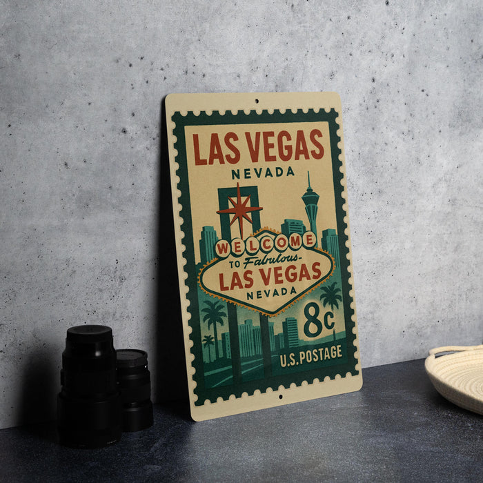 A vintage-style metal sign with the text "LAS VEGAS NEVADA" and "WELCOME TO FAVOROUS LAS VEGAS NEVADA" printed on it, along with a star and a city skyline. The sign is placed on a gray surface, and there are some black camera lenses and a white dish visible in the background.