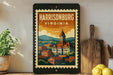 A framed vintage postage stamp depicting a cityscape of Harrisonburg, Virginia, with a clock tower and buildings, is displayed on a shelf next to a wooden cutting board and a potted plant.