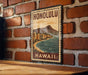 A vintage-style metal sign with the text "HONOLULU" and "HAWAII" printed on it, hanging on a brick wall.