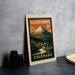 A vintage postage stamp depicting a mountain landscape with a small town in the foreground, placed on a gray countertop next to some black camera lenses.