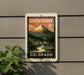 A vintage-style postage stamp from Colorado, featuring a mountain landscape with a winding river and small houses, mounted on a gray wall.