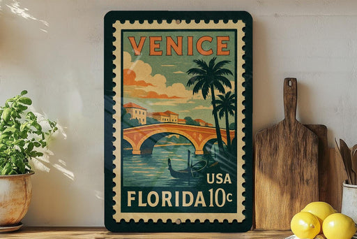 A vintage postage stamp from Venice, Florida, featuring a scenic illustration of a bridge and palm trees, is displayed on a shelf next to a wooden cutting board and a potted plant.