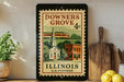 A vintage postage stamp from Illinois, featuring a church and a train, is displayed on a shelf next to a wooden cutting board and a potted plant.