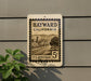 A vintage-style postage stamp from the United States, featuring an illustration of a town in California with a road and buildings, mounted on a wall.