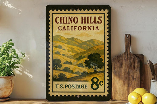 A framed vintage postage stamp from the United States, depicting a scenic landscape of hills and mountains, is displayed on a shelf next to a wooden cutting board and some potted plants.
