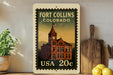 A framed vintage-style stamp depicting the Fort Collins, Colorado, United States, with a clock tower and a building, placed on a shelf next to a wooden cutting board and some lemons.